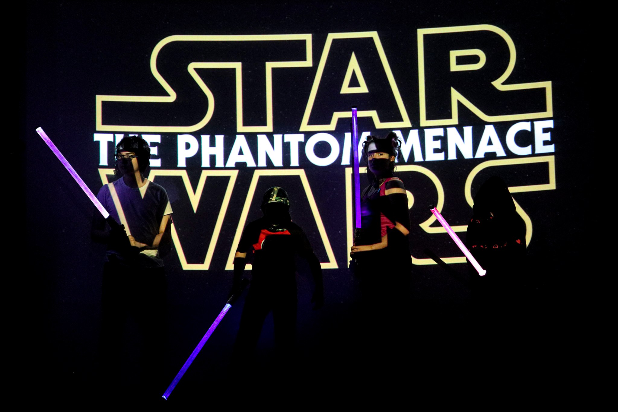 4 tweens stand in front of an illuminated logo of Star Wars Episode 1 while brandishing lightsabers in a defensive position