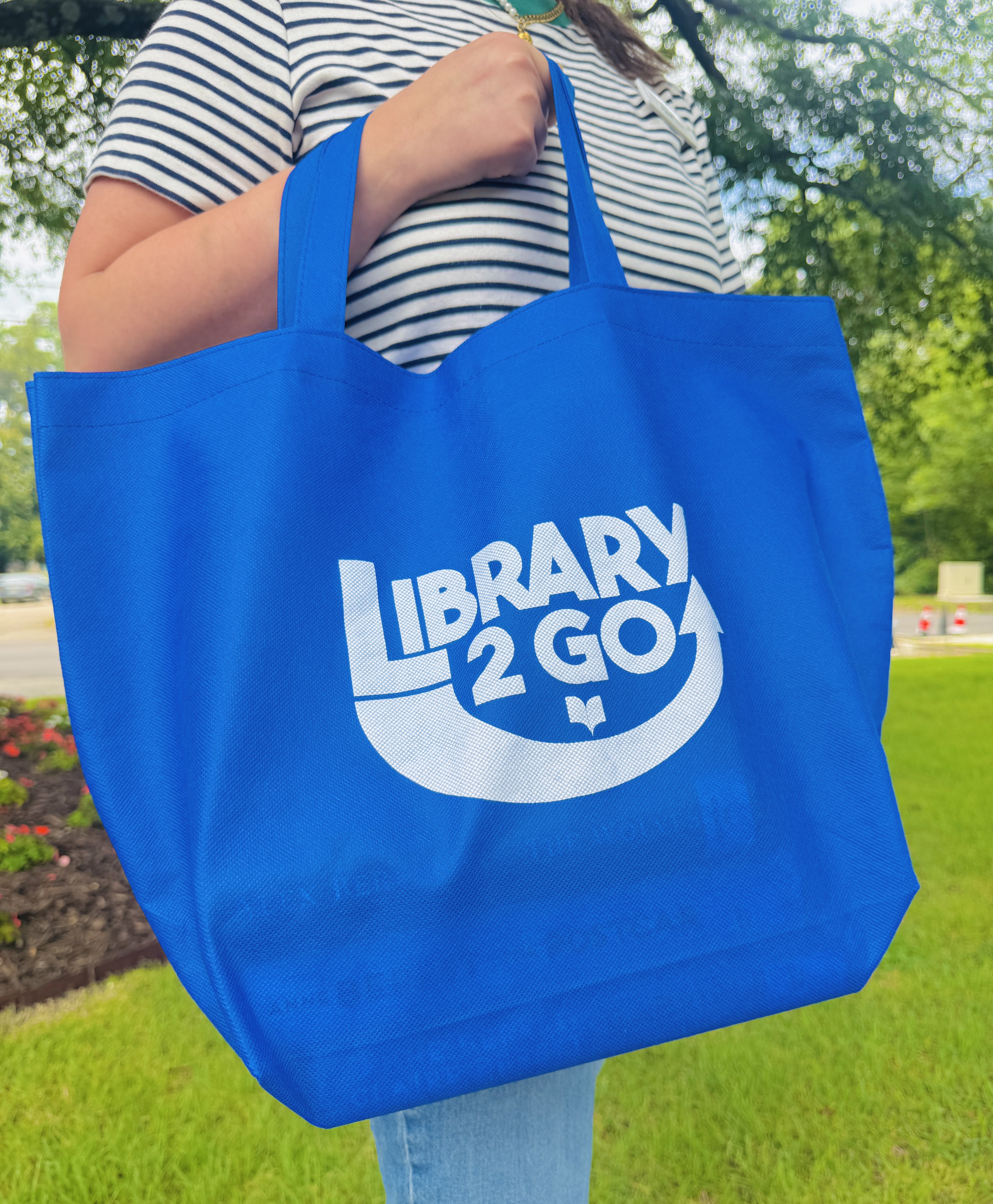 A person whose face is not in view stands in grass and holds a blue bag that says "Library 2 Go"