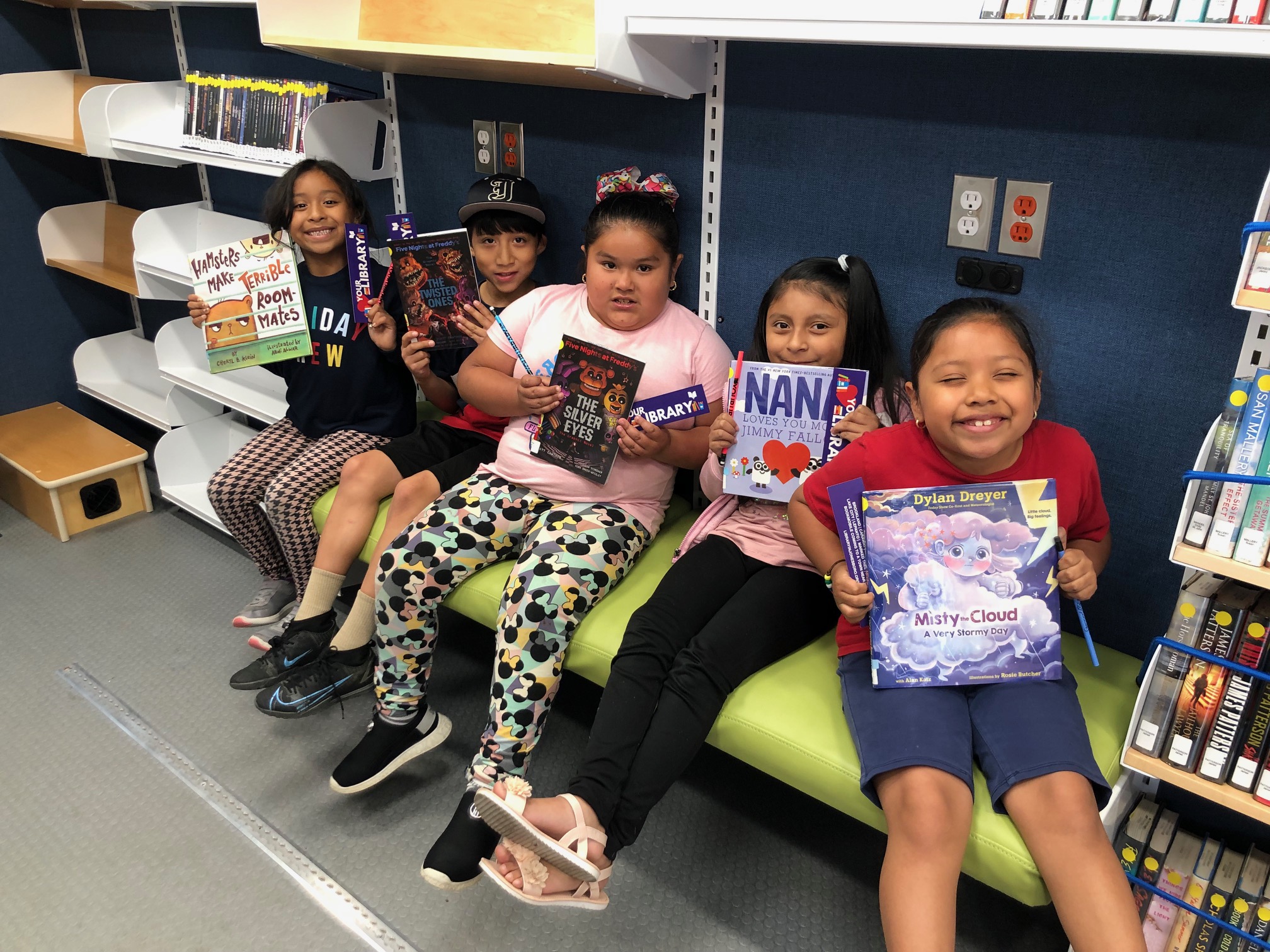 5 young girls sit on the bench inside the bookmobile, showing off the books they checked out