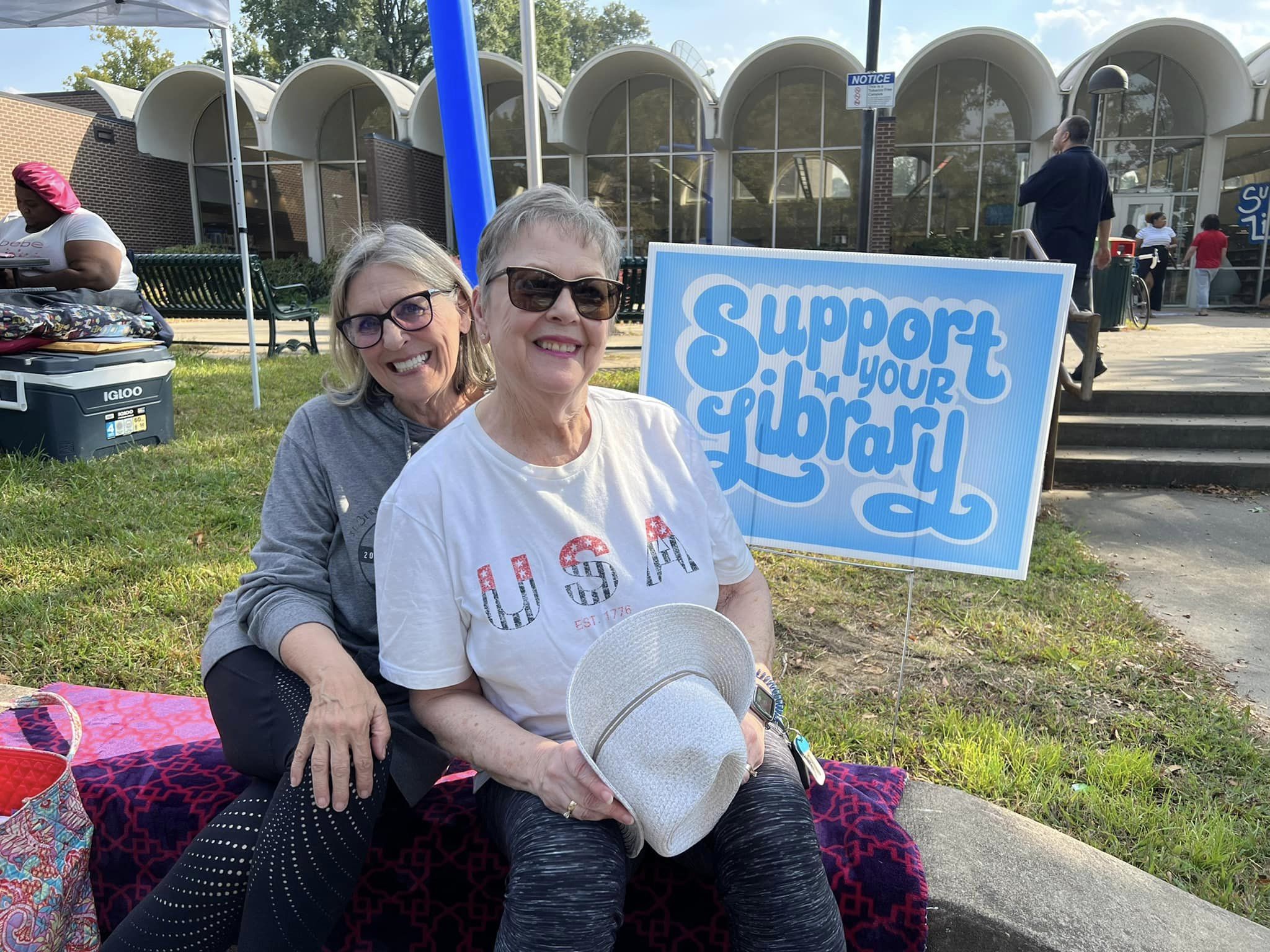 Two women on the CCJPL lawn smile and pose next to a Support Your Library yard sign