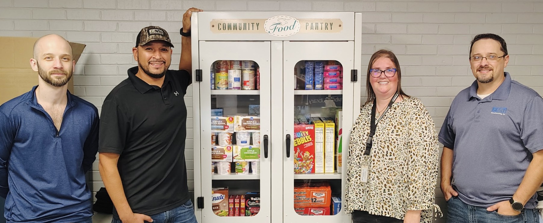 Four Best Manufacturing, Inc. employees stand on either side of their newly donated food pantry, which is completely stocked with nonperishables.